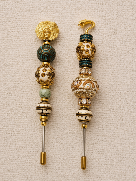 Pair of ivory, teal, and gold beaded cigar nubbers with a medallion face topper on one piece and a serpent topper on the other, displayed on a soft beige background.
