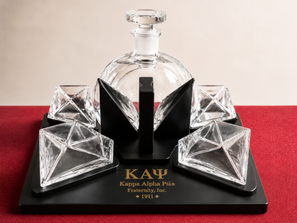 Faceted glass decanter set with four angular tumblers on a black Kappa Alpha Psi display stand against a deep red surface.
